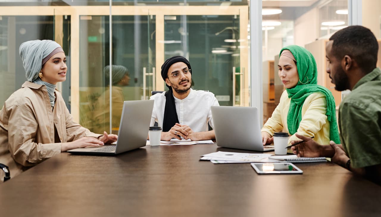 أربعة زملاء يتحدثون حول طاولة اجتماعات عليها حواسيب محمولة