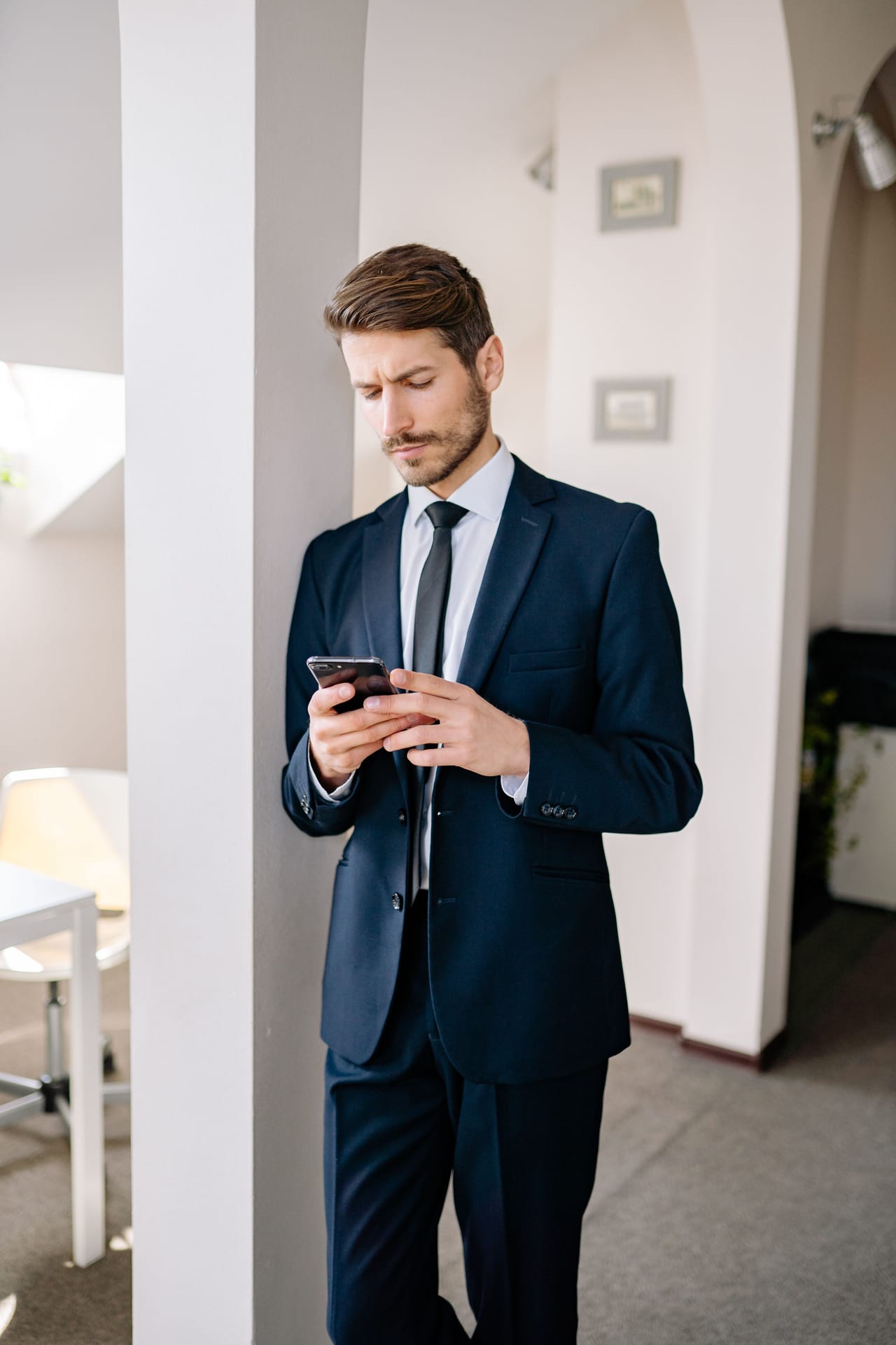 A man in a navy suit leaning against a pillar as he reads his phone